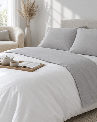 Light Grey plain cotton flat sheet turned down over a white duvet with matching pillows, a tea tray and boucle armchair.