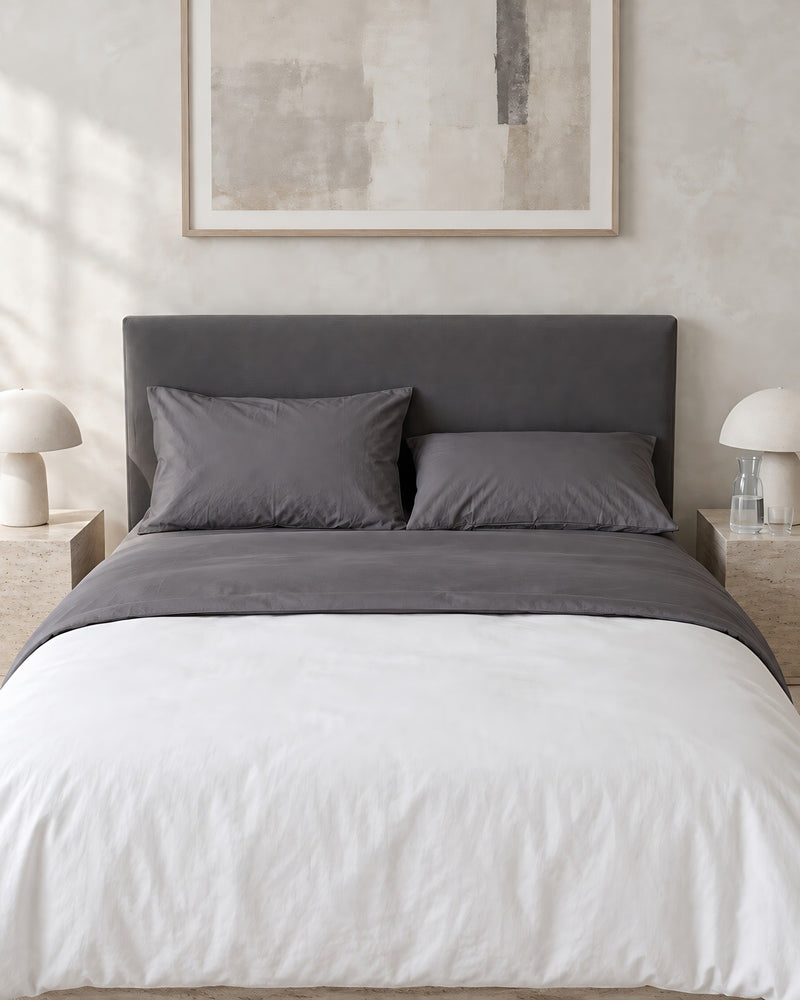 Charcoal Grey plain cotton flat sheet against a limewashed plaster wall between travertine bedside tables