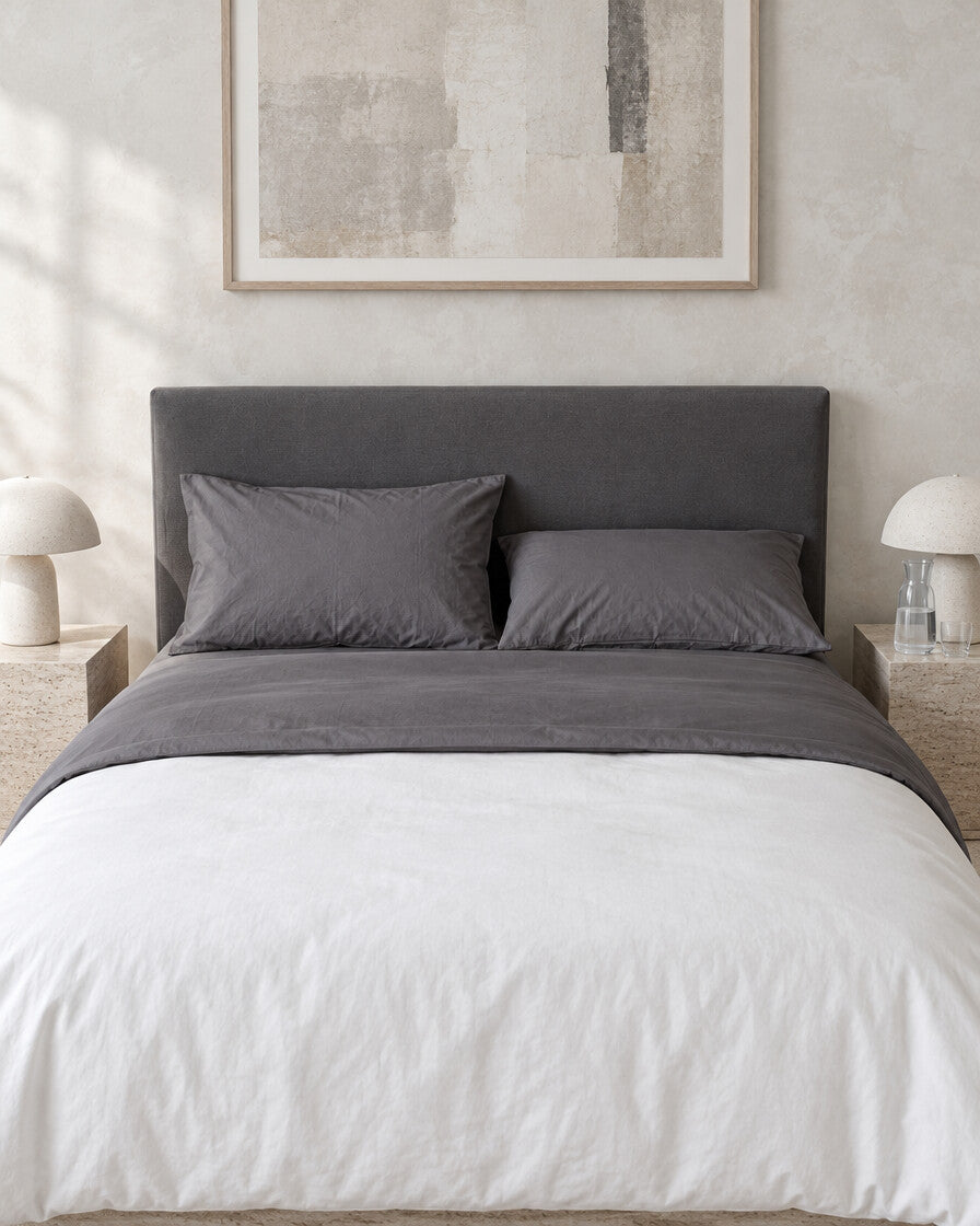 Charcoal grey plain cotton flat sheet against a limewashed plaster wall between travertine bedside tables