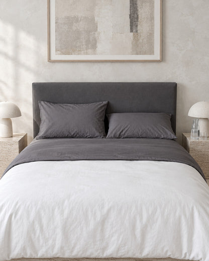 Charcoal grey plain cotton flat sheet against a limewashed plaster wall between travertine bedside tables