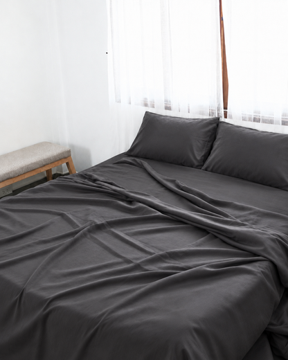 Charcoal Grey silky cotton flat sheet, a grey cotton sateen flat sheet, with matching pillowcases on a bed beside a bright window.