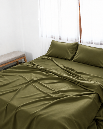Juniper Green silky cotton flat sheet, an olive green cotton sateen flat sheet, with matching pillowcases on a bed beside a bright window.