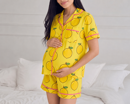 A pregnant woman wearing a yellow short-sleeve pajama set with a lemon print and pink trim, standing in front of a white bed.