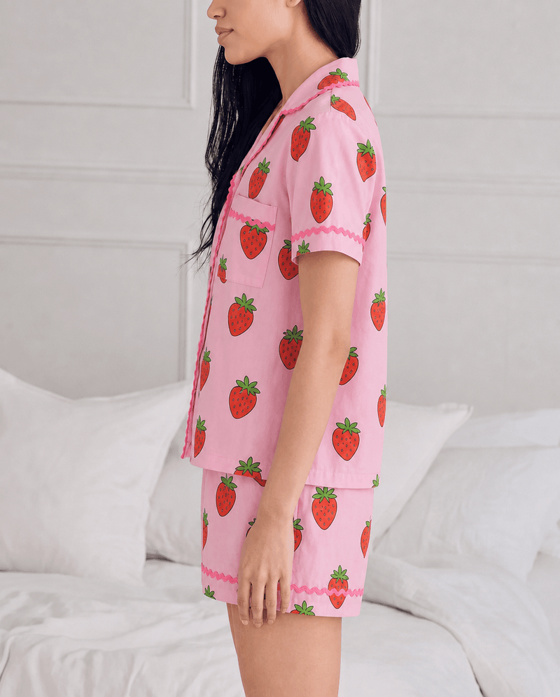 A woman wearing a pink pajama set with a strawberry print, standing in a bedroom setting with white bedding and a white paneled wall behind her.