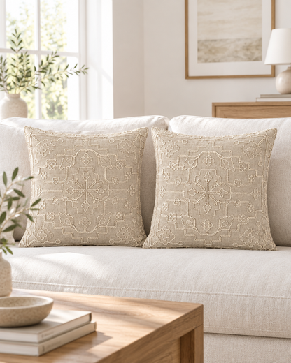 Two oat ramie cotton cushions with a symmetric cream cord-embroidered medallion on a linen sofa in a sunlit Scandi room