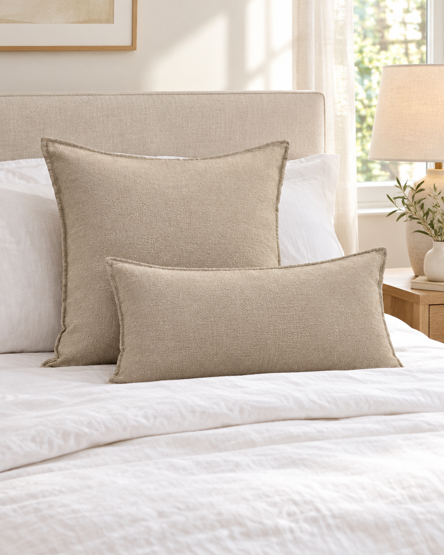 Plain oat slub linen cotton cushions, square and lumbar, on a made bed with white linen and a bedside lamp