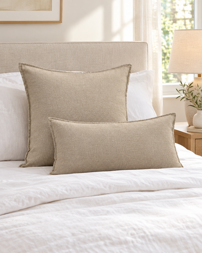 Plain oat slub linen cotton cushions, square and lumbar, on a made bed with white linen and a bedside lamp