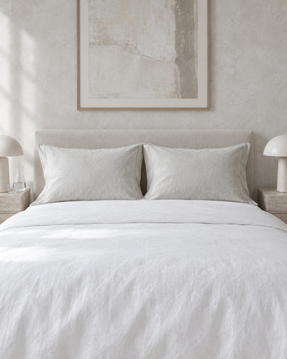 White French linen pillowcases against a limewashed plaster wall between travertine bedside tables