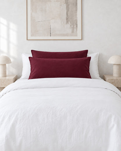 Burgundy Red plain cotton pillowcases on pillows against a white bed between bedside lamps