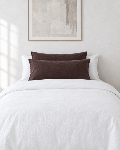 Chocolate plain cotton pillowcases on pillows against a white bed between bedside lamps.