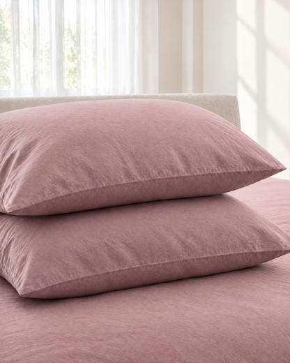Two Dust Pink plain cotton pillowcases stacked on a matching sheet before a linen headboard