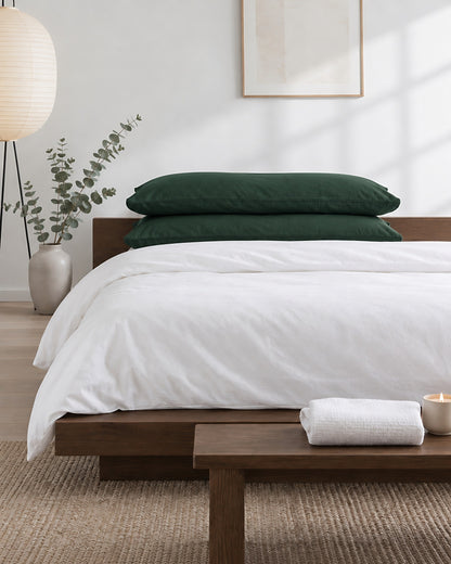 Forest Green plain cotton pillowcases in bottle green stacked on a low oak bed with a white duvet