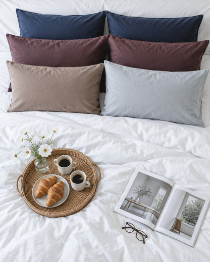 Navy, burgundy, taupe and grey plain cotton pillowcases seen from above with a breakfast tray of coffee and croissants