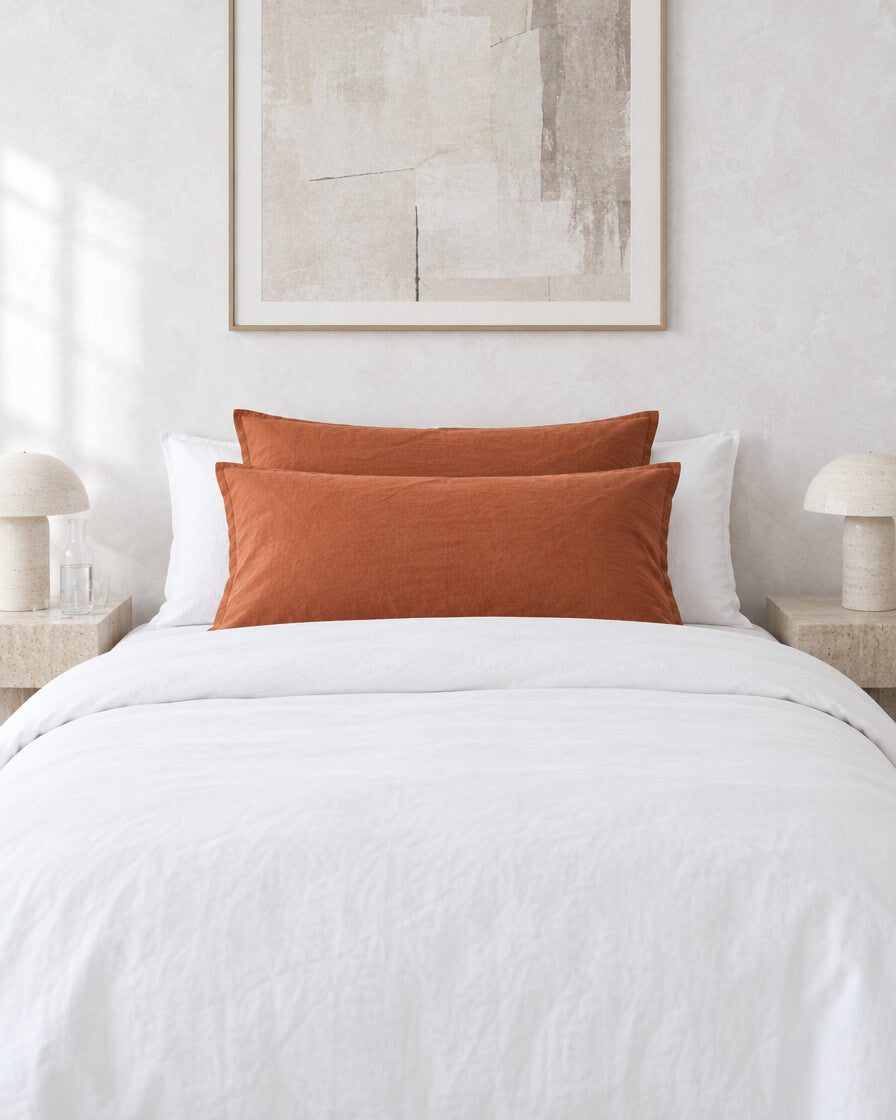 Burnt orange plain cotton pillowcases against a limewashed plaster wall between travertine bedside tables