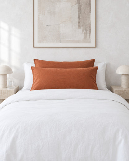 Burnt orange plain cotton pillowcases against a limewashed plaster wall between travertine bedside tables