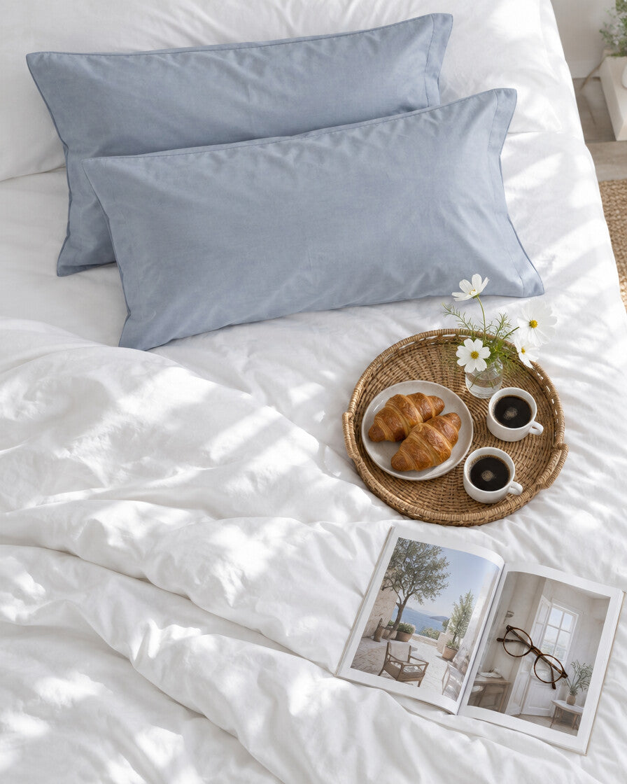 Blue-grey plain cotton pillowcases seen from above with a breakfast tray of coffee and croissants