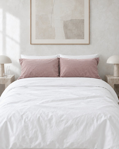 Dusty pink plain cotton pillowcases against a limewashed plaster wall between travertine bedside tables