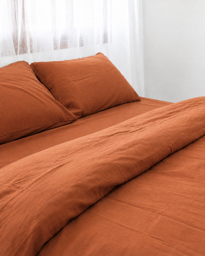 Two pillows in Terracotta plain cotton pillowcases at the head of a made bed by a white sheer curtain.