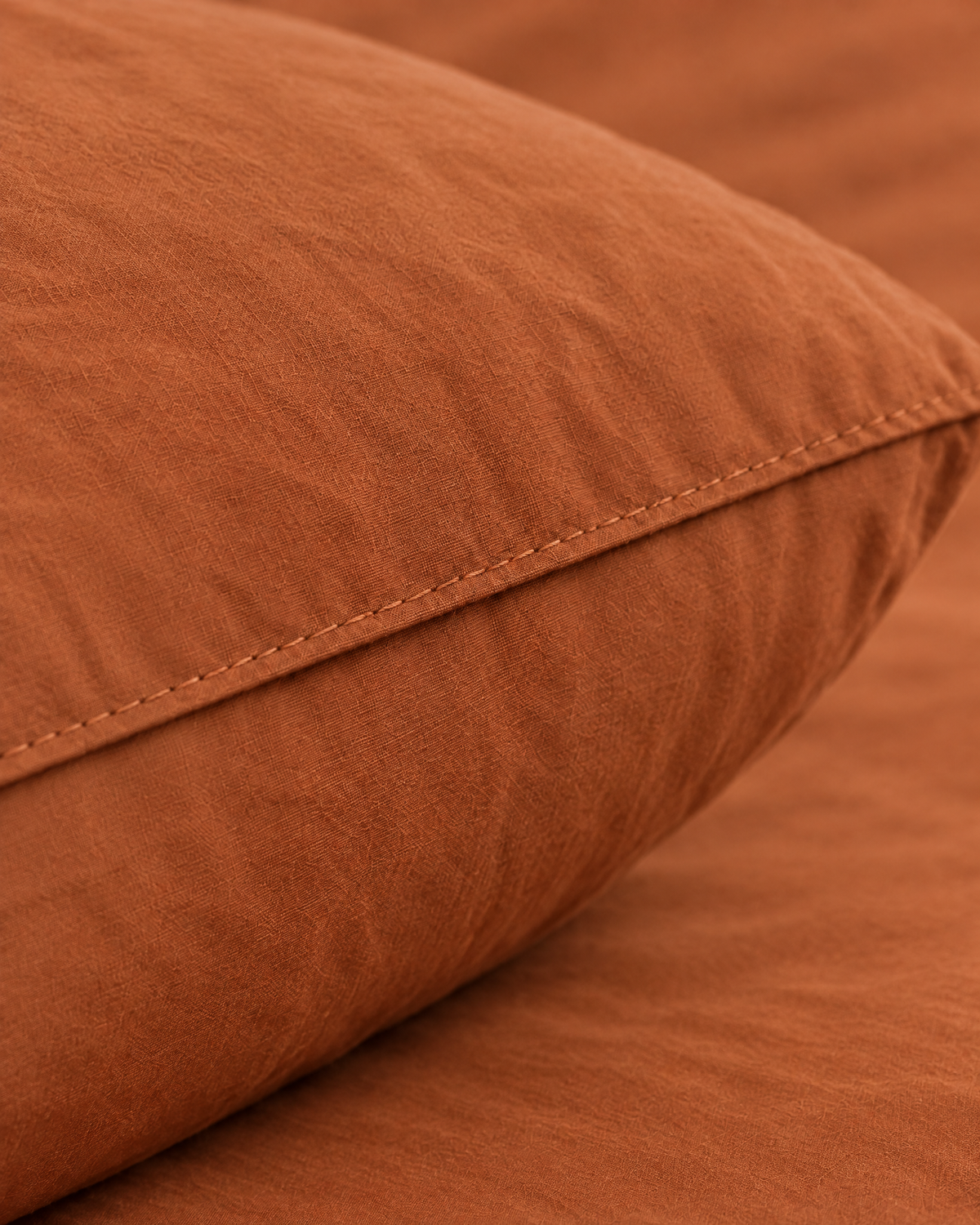 Macro of the stitched edge of a Terracotta plain cotton pillowcase on a plump pillow, showing the washed cotton weave.