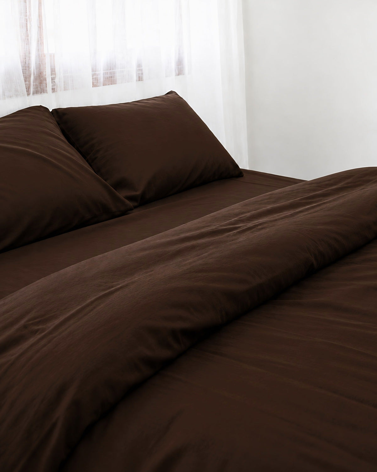 Two pillows in Chocolate silky cotton pillowcases at the head of a made bed by a sheer curtain