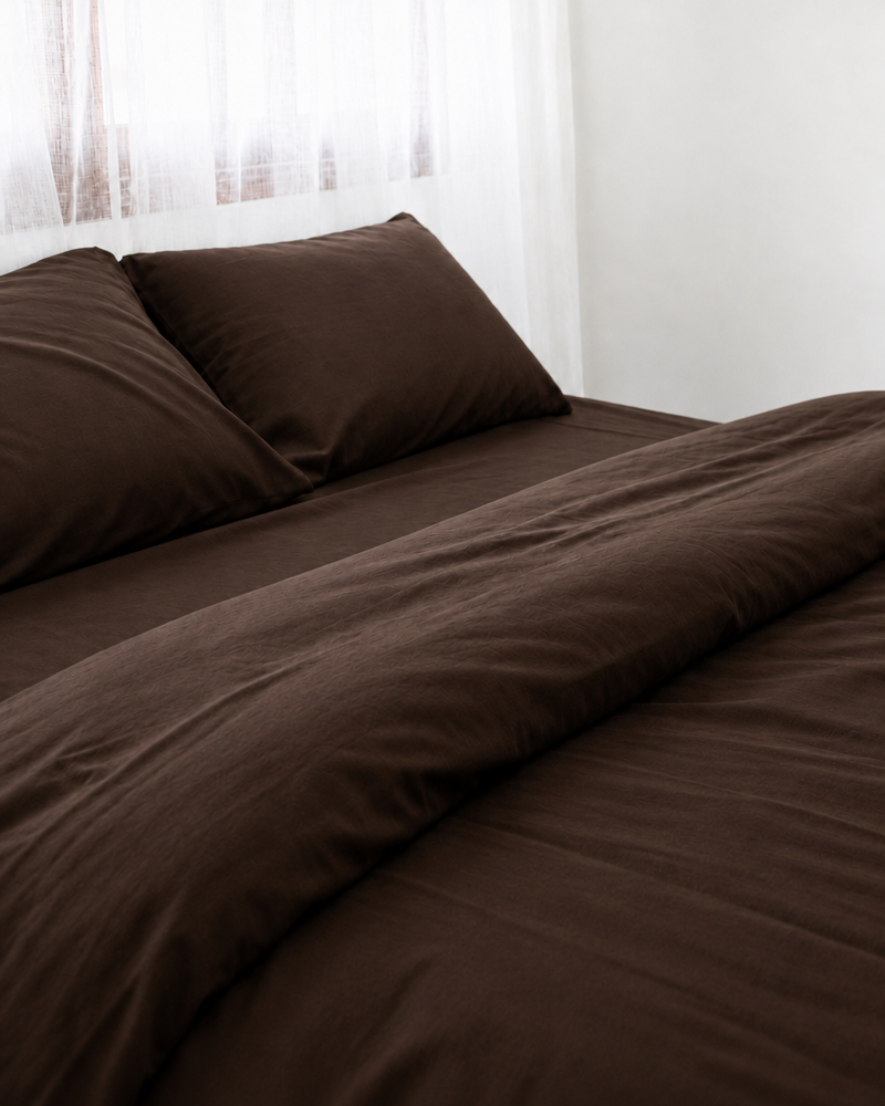 Two pillows in Chocolate silky cotton pillowcases at the head of a made bed by a sheer curtain.