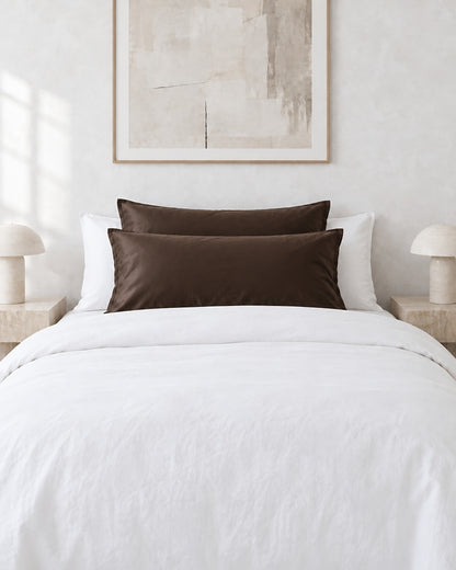 Chocolate silky cotton pillowcases on pillows against a white bed between bedside lamps