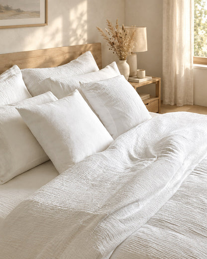 White cloud cotton pillows and a rumpled duvet, styled in a calm sunlit Scandinavian bedroom
