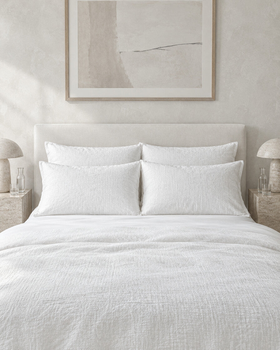 White cloud cotton pillowcases against a limewashed plaster wall between travertine bedside tables