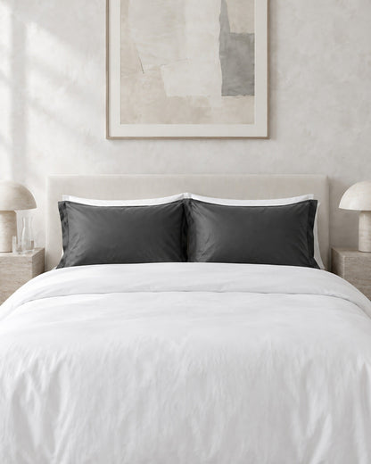 Charcoal Grey silky cotton pillowcases against a limewashed plaster wall between travertine bedside tables