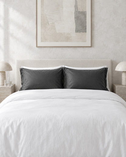 Charcoal grey silky cotton pillowcases against a limewashed plaster wall between travertine bedside tables