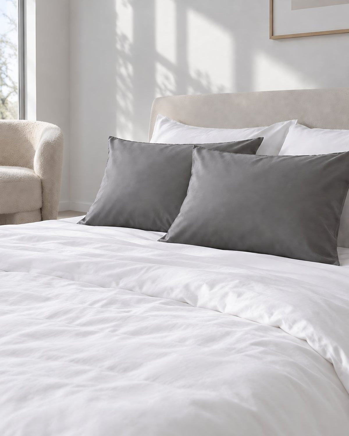 Single grey silky cotton pillowcases on a white bed, a boucle armchair in the corner