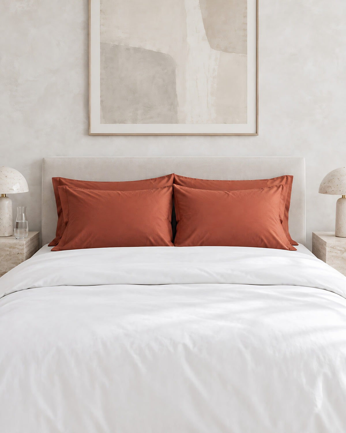 Rust silky cotton pillowcases against a limewashed plaster wall between travertine bedside tables