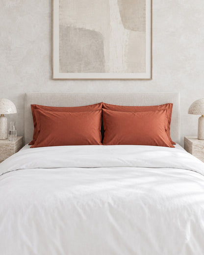 Terracotta silky cotton pillowcases against a limewashed plaster wall between travertine bedside tables