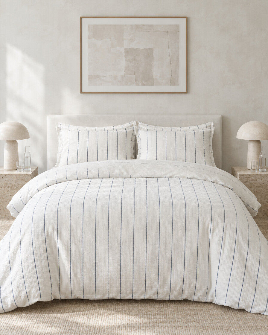 Cream pinstripe cloud cotton bedding set against a limewashed plaster wall between travertine bedside tables