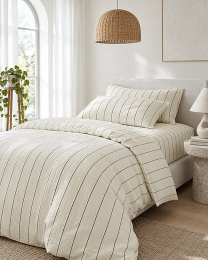 Cream pinstripe cloud cotton bedding set beneath an arched window with a trailing pothos and rattan pendant