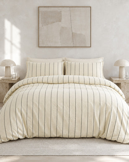 Cream pinstripe cloud cotton bedding set against a limewashed plaster wall between travertine bedside tables, styled in calm neutrals
