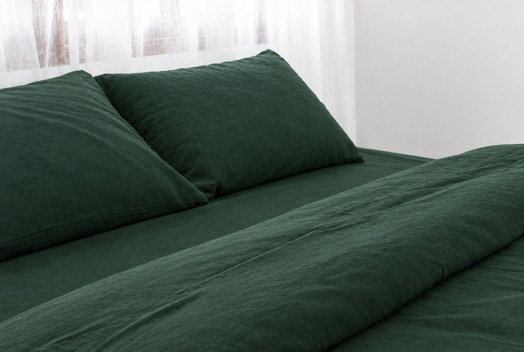 Two pillows in Forest Green plain cotton pillowcases at the head of a made bed with a matching bottle-green sheet and duvet cover