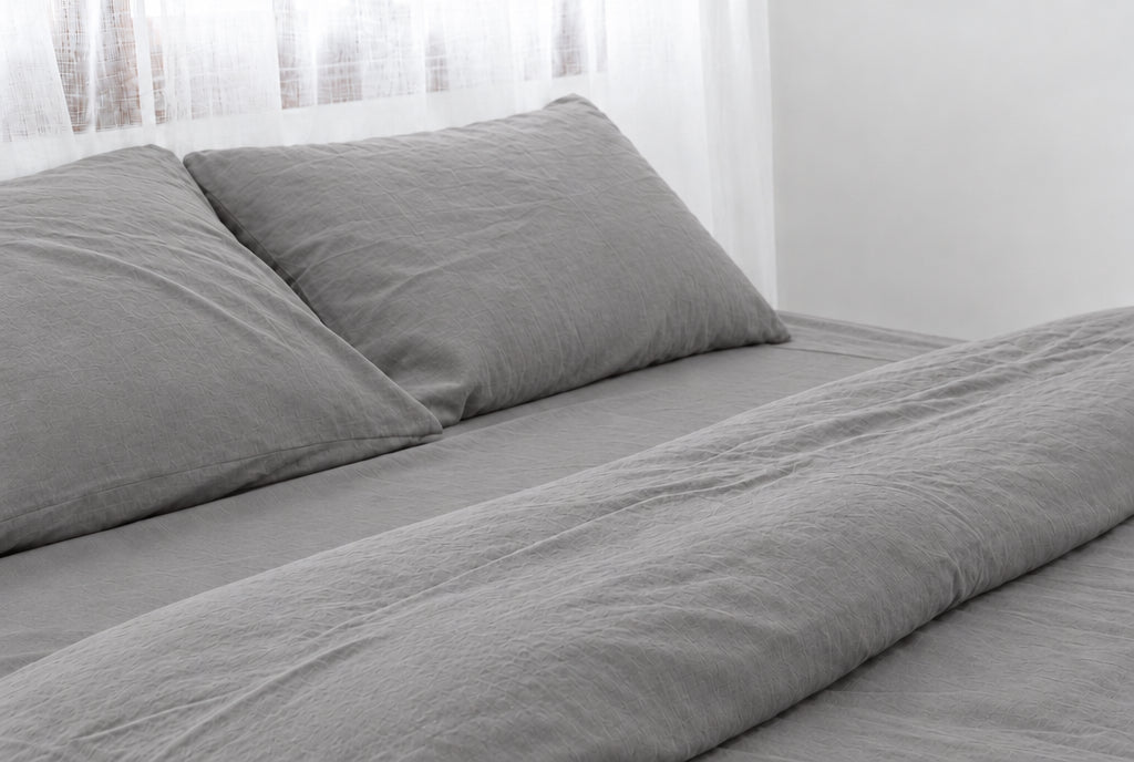 Two pillows in Smoke plain cotton pillowcases at the head of a made bed with a matching Smoke grey percale sheet and duvet, horizontal close-up