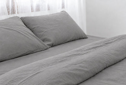Two pillows in Smoke plain cotton pillowcases at the head of a made bed with a matching Smoke grey percale sheet and duvet, horizontal close-up