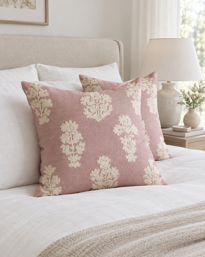 Rose pink cushions with ivory floral embroidery on a made bed with white linen and a bedside lamp
