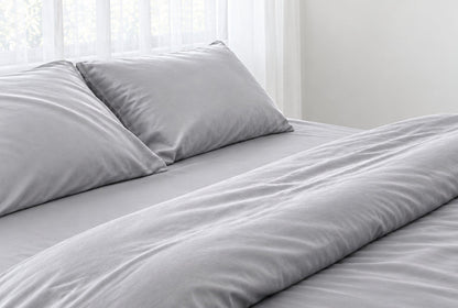 Two pillows in Silver silky cotton sateen pillowcases at the head of a made bed with a matching sheet and duvet, horizontal close-up