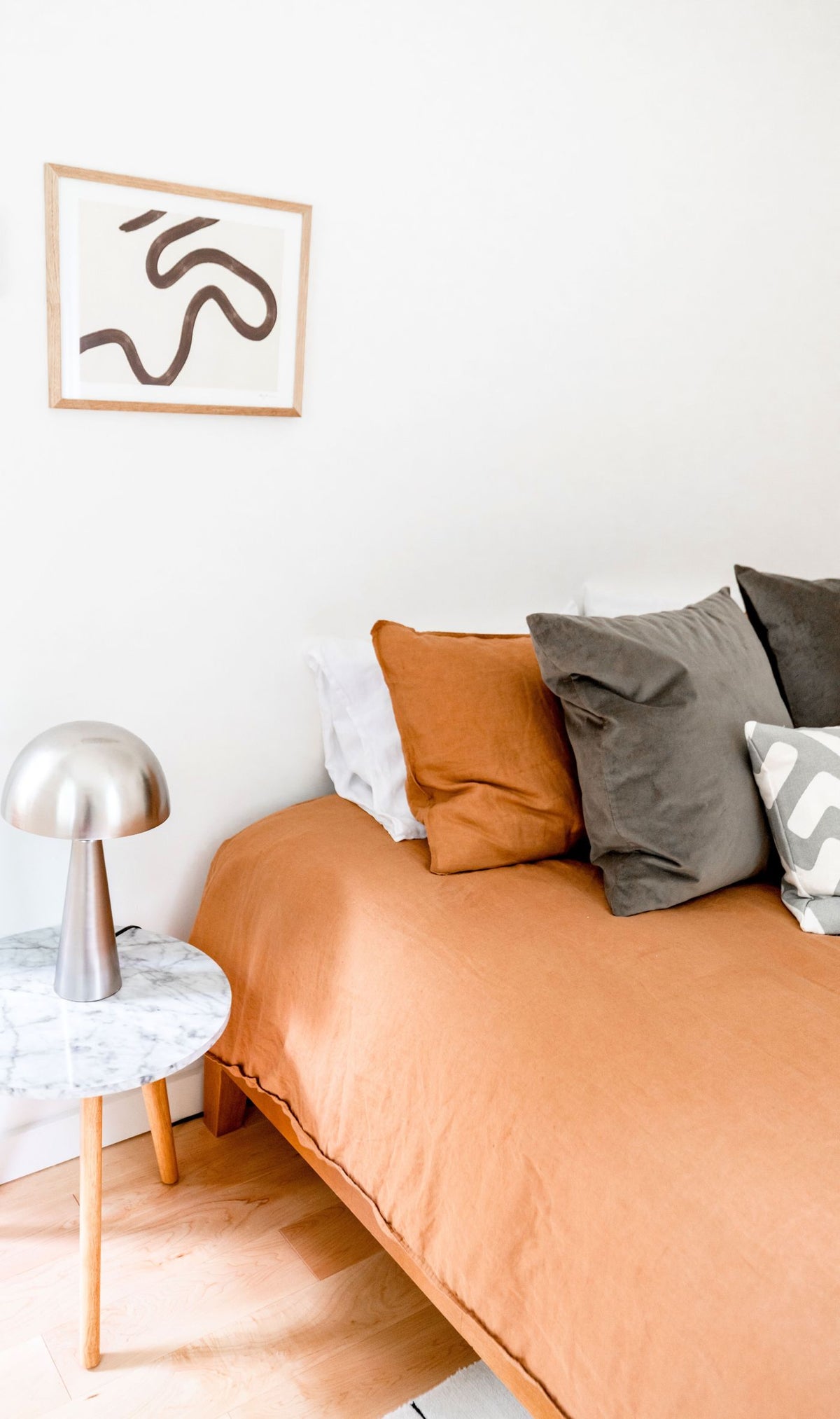 A bed dressed in a terracotta duvet cover and pillow, white wall and oak floor
