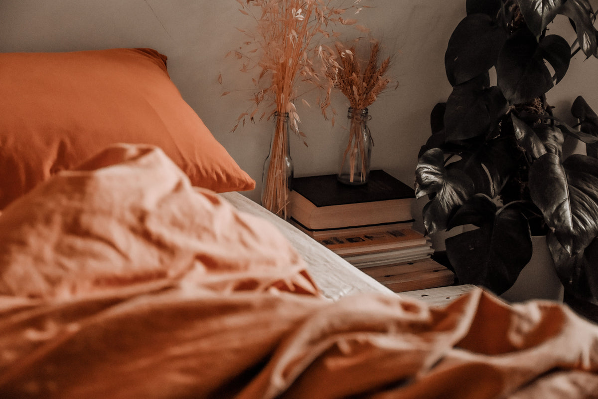 An unmade bed in creased terracotta-orange bedding, dried grasses on the bedside table