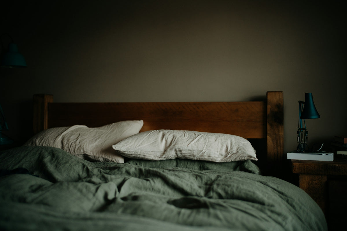 A wooden bed in olive green bedding with pale pillows, lamp on the side table