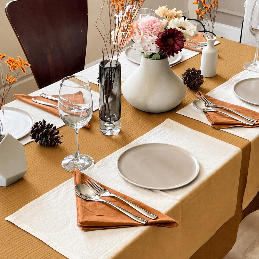 Mustard cloud cotton tablecloth dressed for dinner with grey plates, wine glasses and a vase of dried flowers.