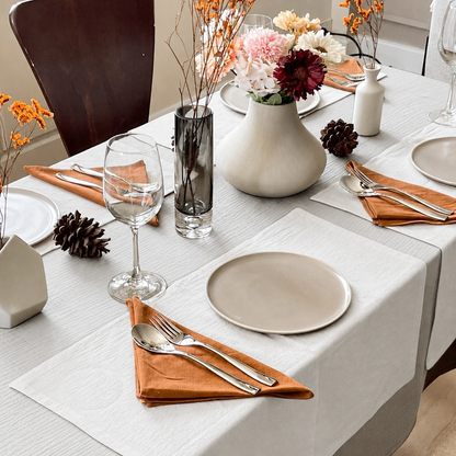Off White cloud cotton tablecloth under a cream runner, set with orange napkins, dried flowers and pinecones.