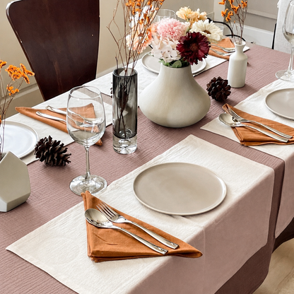 Pale Pink cloud cotton tablecloth with a cream runner, white vase of dried blooms and orange napkins.