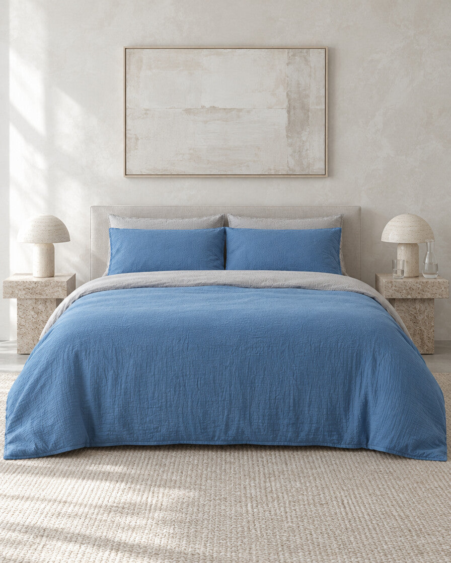 Blue and sand two-tone cloud cotton bedding against a limewashed plaster wall between travertine bedside tables