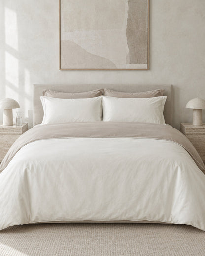 Cream two-tone plain cotton bedding against a limewashed plaster wall between travertine bedside tables